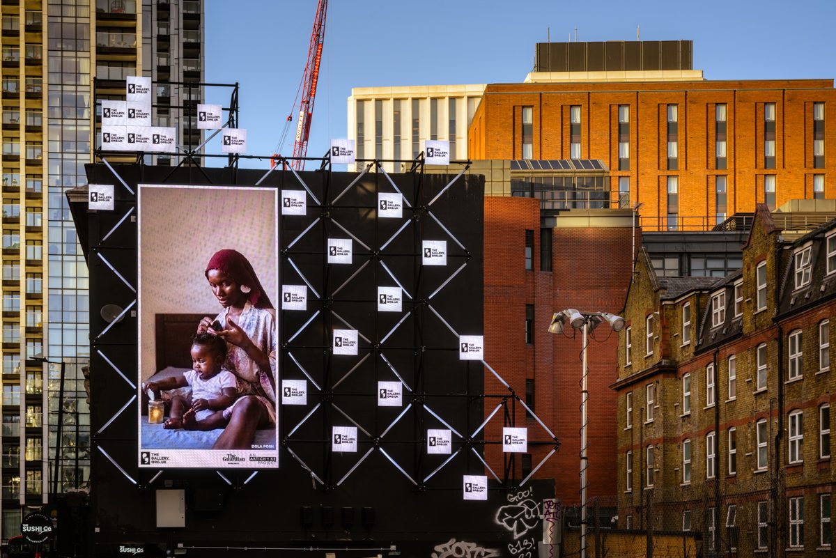 ‘Care’ (2021) by Dola Posh. The Gallery, Season 2, 2023. Produced by Artichoke. Photo by Yves Salmon. Old Street Digital Canvas, London. Landscape 2 SMALL A billboard of the artwork 'Care' by Dola Posh on a busy London road. A black woman sits atop her bed with a toddler sat cross-legged in front of her. She is wearing a floral robe with a burgundy head wrap. The woman combs out her daughter's afro and puts it into twists.