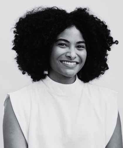 A b&w image of a young POC woman with large afro hair. She is smiling into the camera and wears a white turtleneck top.