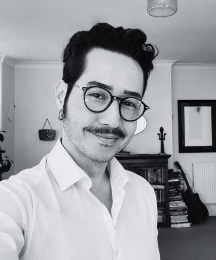 A b&w selfie of a man wearing a white shirt. He is smiling into the camera. He wears dark-rimmed round glasses and has a moustache and short hair.