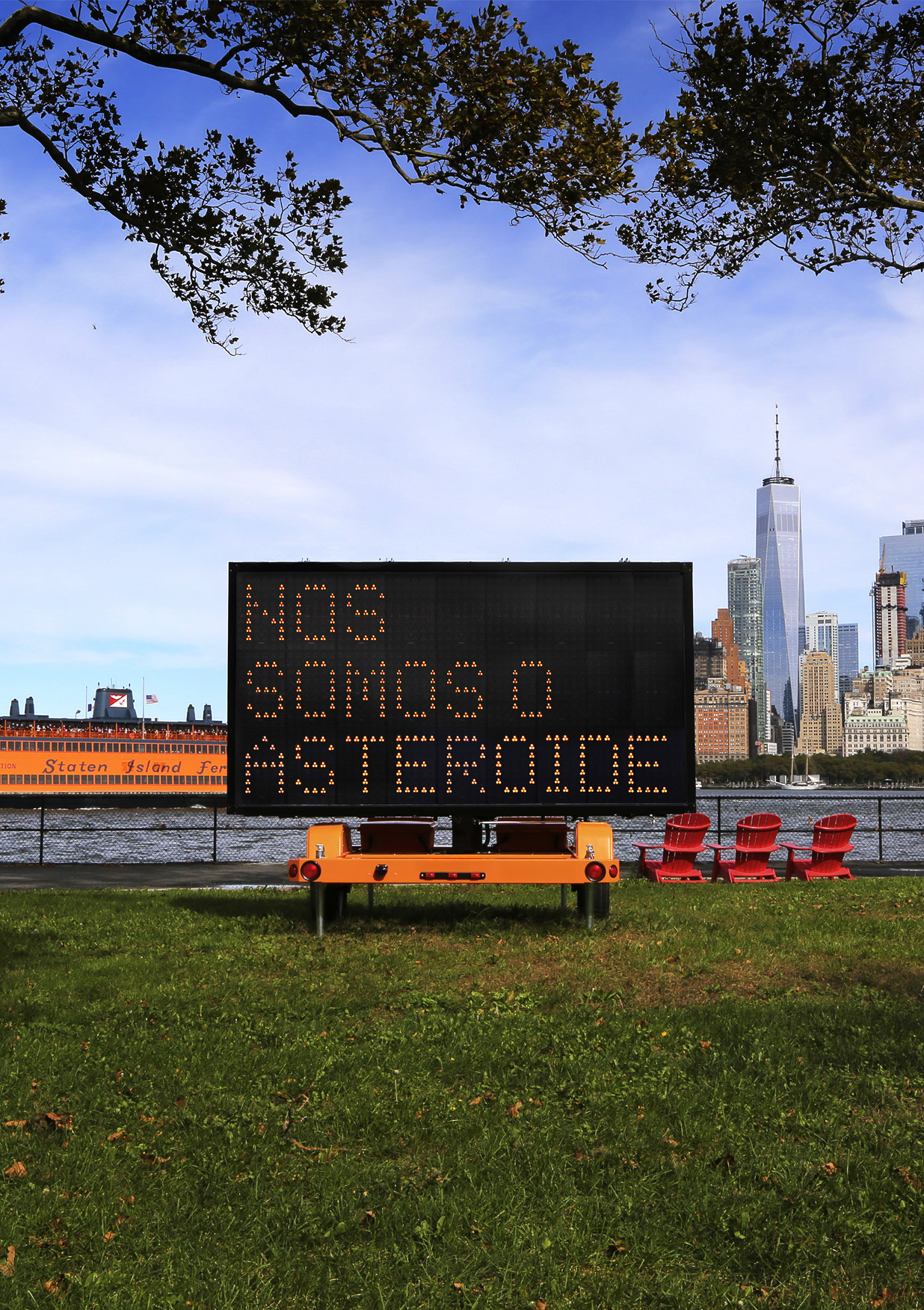 A digital sign reading "WE ARE THE ASTEROID" is set on grass near a river. Background shows a Staten Island Ferry and New York City skyline. The image is split into 3 parts, the foreground is grass, the middle sliver is the river and the background is the sky.