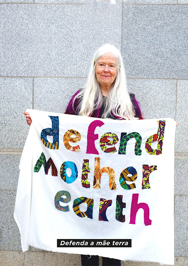 An older woman with long white hair holds a colourful banner reading "defend mother earth" against a grey wall. The banner is white cloth with patchwork lettering created from various scraps of patterned fabric.