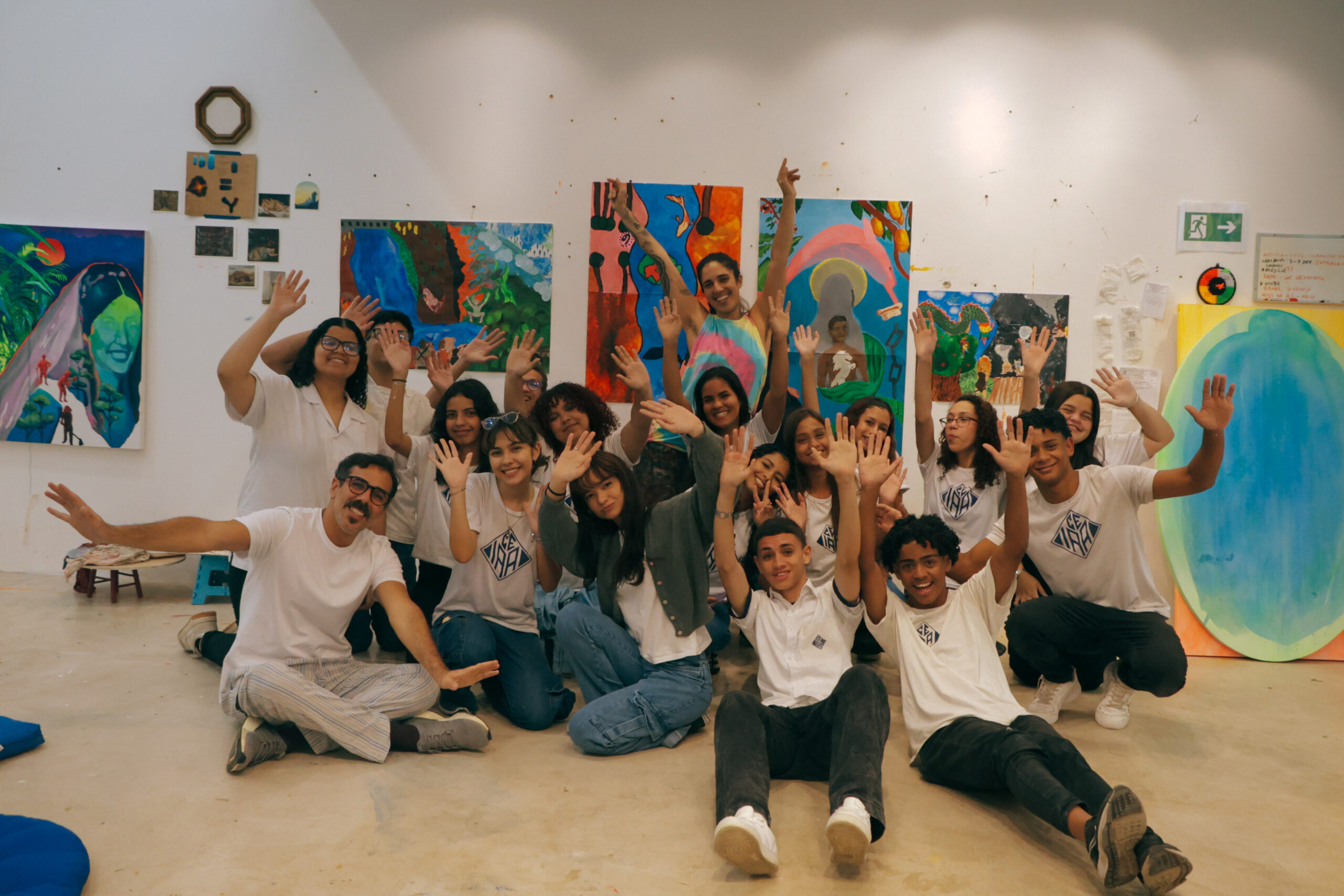 A joyful group of young people in a bright art studio in Brazil. They are wearing casual outfits and raising their hands in celebration. Colourful paintings adorn the walls behind them.