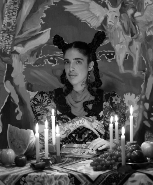 A person looks into the camera. They are wearing their dark hair in plaits and their style is very Frida-esque. They are sat at a table dressed with candelabras and fruit and there is something very mystical about the image.