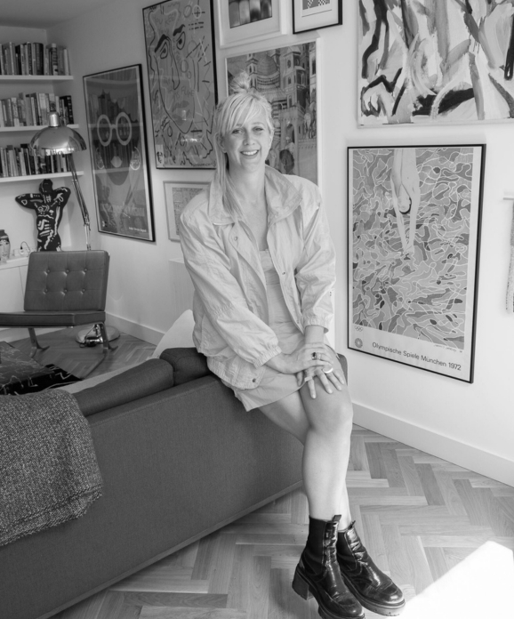 A B&W image of a woman with short blonde hair perching on a sofa. She is smiling into the camera and has her hands on her crossed legs. There are several paintings hung up on the wall behind her.