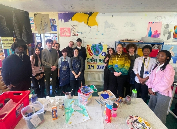 A group of about fifteen students and staff members pose together in a bright, busy art classroom. They are surrounded by art supplies, including paint containers and brushes on a central table, with various student sketches and colourful murals displayed on the walls behind them.
