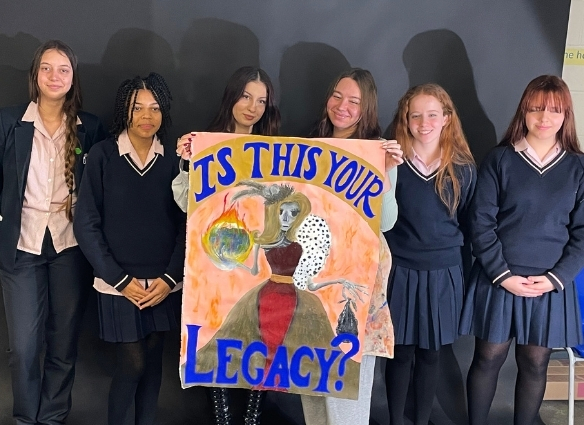 Six students stand in a row against a dark backdrop, holding up a large, hand-painted poster. The artwork depicts a skeletal figure in a gown holding a flaming Earth, with the bold blue text "IS THIS YOUR LEGACY?" at the top and bottom.